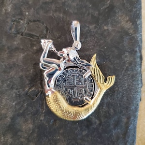 May include: A silver and gold mermaid pendant featuring a silver coin with a cross design. The pendant has a loop at the top for attaching to a chain.