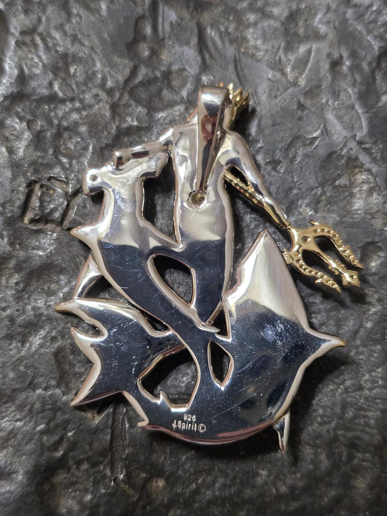 May include: A silver-toned pendant featuring a stylized depiction of a merman holding a trident. The merman is adorned with a crown and is surrounded by a stylized aquatic design. The pendant is marked with "925" and "Spirit".