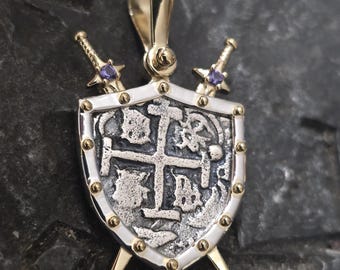 Puede incluir: Pendiente de plata y oro con un escudo con un diseño de cruz y dos espadas con piedras preciosas púrpuras.