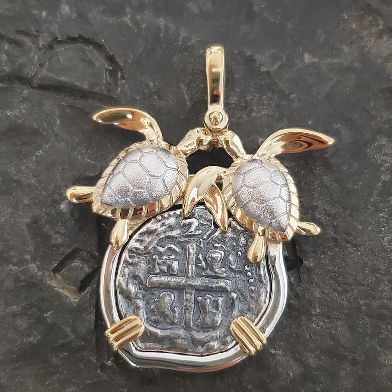 May include: A two-tone pendant featuring a silver coin with a cross design, framed in silver and gold. Two gold and silver turtles flank the coin, with a gold bail at the top. The pendant is set against a dark, textured background.