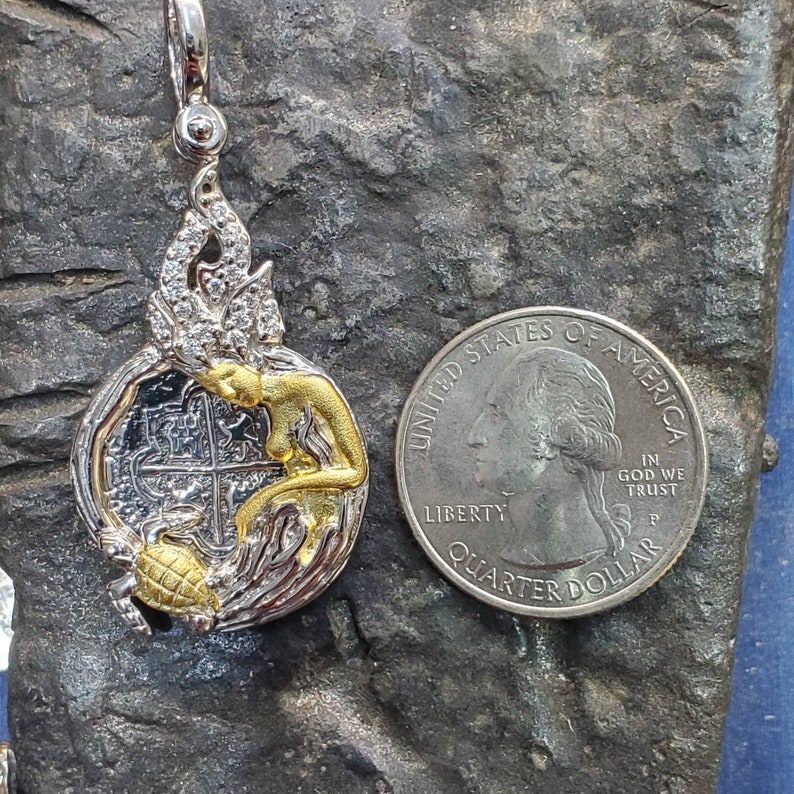 May include: Silver and gold pendant featuring a mermaid with a treasure map inside a circle. The pendant is about the size of a US quarter.