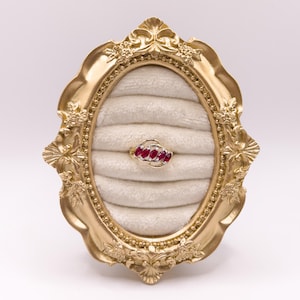 May include: A gold ring featuring five marquise-cut red rubies arranged in a diagonal row, set within a polished gold band. The ring is displayed in an oval, gold-toned frame with an ornate, floral-embossed border and a cream-colored velvet insert.
