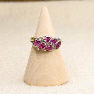 May include: A gold ring featuring a cluster of six marquise-cut magenta gemstones arranged in a diagonal pattern. The stones are set in a silver-toned metal mounting that contrasts with the gold band, creating a distinct visual focal point.