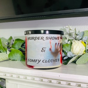 May include: A white candle with a red and white design. The candle has a black lid and the text "Murder Shows & Comfy Clothes" printed on the front.