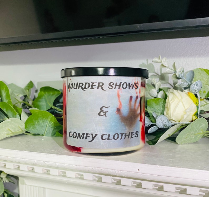 May include: A white candle with a red and white design. The candle has a black lid and the text "Murder Shows & Comfy Clothes" printed on the front.