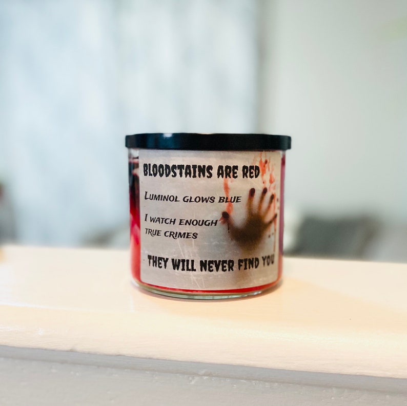 May include: A red candle with a black lid and a white label that reads "BLOODSTAINS ARE RED, LUMINOL GLOWS BLUE, I WATCH ENOUGH TRUE CRIMES, THEY WILL NEVER FIND YOU".