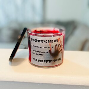 May include: A red candle with a white label that reads "BLOODSTAINS ARE RED, LUMINOL GLOWS BLUE, I WATCH ENOUGH TRUE CRIMES, THEY WILL NEVER FIND YOU". The label also features a bloody handprint.