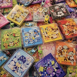 May include: A collection of square acrylic keychains featuring colourful illustrations of various cartoon creatures. Each keychain displays a group of characters in a specific colour theme, including red, blue, green, yellow, and purple, with attached metal clasps.