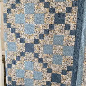 May include: A patchwork quilt featuring a repeating pattern of squares in shades of light blue, dark navy blue, and a floral print with yellow and white blossoms. The design consists of alternating solid-coloured blocks and intricate floral fabric.