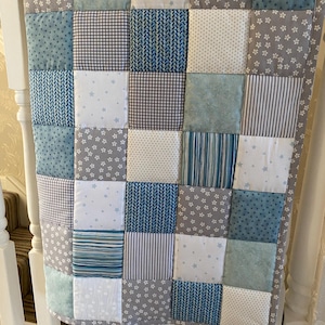 May include: A patchwork quilt in blue, grey, and white. The quilt features a variety of patterns, including stars, stripes, and floral motifs. The design is made from square fabric pieces, creating a textured and visually appealing design. The quilt is likely for a baby or child.