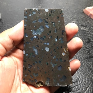 Pode incluir: Uma placa retangular de pedra polida com uma cor de base marrom escuro. A pedra apresenta padrões irregulares de inclusões azuis, brancas e verdes. A pedra é segurada em uma mão.