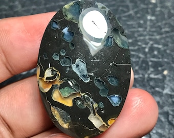Pode incluir: Uma pedra preciosa oval com um fundo escuro e salpicado. A pedra apresenta uma grande área branca e manchas de azul, verde e amarelo. A pedra preciosa é segurada em uma mão, mostrando sua superfície polida e variações de cores únicas.