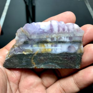 May include: A slab of purple, white, and yellow banded agate with a dark gray base. The agate has a layered appearance with a smooth, polished surface.