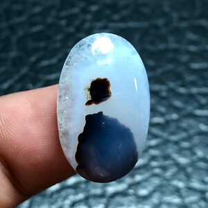 May include: A white oval cabochon with a dark grey and black agate pattern. The agate has a cloudy, translucent appearance.