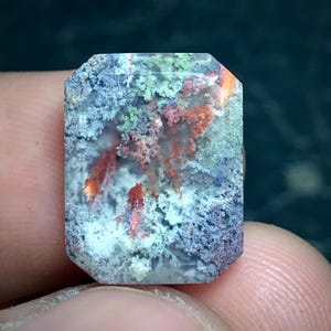 May include: A rectangular, faceted gemstone featuring a translucent, milky white base with intricate, moss-like inclusions in shades of vibrant orange, deep forest green, and muted grey, creating a complex, organic pattern throughout the stone.