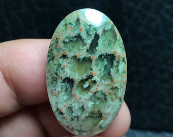 May include: A green and orange agate cabochon with a unique pattern of white and orange inclusions. The cabochon is oval shaped and has a smooth, polished surface.