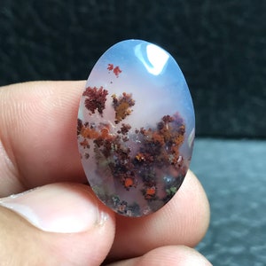May include: A white agate cabochon with red and brown dendritic inclusions. The cabochon is oval shaped and has a smooth, polished surface.