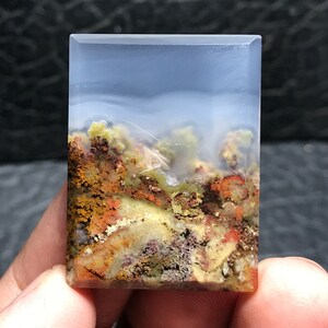 May include: A polished agate cabochon with a landscape scene. The agate is translucent with a white, blue, and gray background. The landscape is made up of brown, orange, yellow, and green colors.