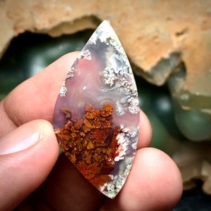 May include: A teardrop-shaped agate cabochon with a white, pink, and brown color scheme. The agate has a dendritic pattern with a white, gray, and brown color scheme.