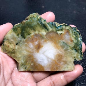 Puede incluir: Una piedra pulida de forma irregular con un patrón natural. La piedra presenta un centro blanco, rodeado de zonas marrones, verdes y amarillas. La piedra se sostiene en una mano.