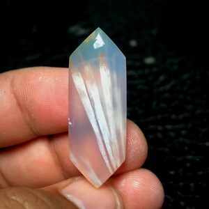 以下が含まれることがあります： 白と薄茶色のインクルージョンが入った、半透明の薄いピンク色のクリスタルポイント。クリスタルは尖った上部と平らな角度の付いた基部を持っています。クリスタルは暗い背景に対して手で持たれています。
