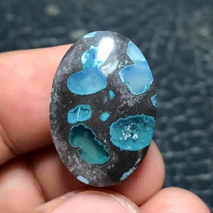 Puede incluir: Una piedra preciosa en forma de cabujón ovalado que presenta una matriz de color gris carbón oscuro con inclusiones incrustadas de color azul turquesa vibrante. La superficie está pulida con un acabado liso y brillante, resaltando el contraste entre la base oscura y las manchas azules irregulares dispersas por la cara de la piedra.