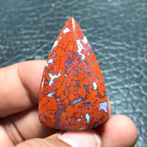 Puede incluir: Una piedra preciosa cabujón de forma triangular pulida que presenta un patrón vibrante de inclusiones de color rojo anaranjado intenso sobre una matriz contrastante de vetas azul claro y gris, con una superficie lisa y brillante que refleja la luz.