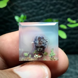 May include: A clear agate cabochon with a dendritic pattern of purple, brown, and green inclusions. The agate is set in a square shape and is approximately 1 inch in size.
