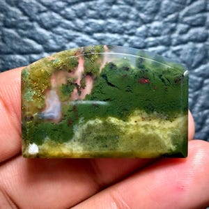 May include: A rectangular, polished gemstone with a rounded top edge. The stone features a landscape-like pattern with shades of green, yellow, and red. The translucent areas reveal a hint of pink and white, creating a unique, natural design.