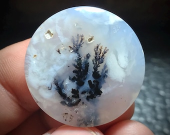 May include: A round, white agate cabochon with black and gray dendritic inclusions. The agate has a cloudy, translucent appearance.