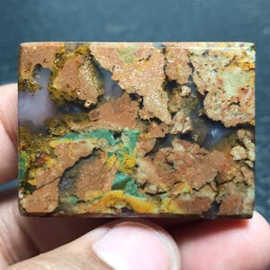 May include: A rectangular polished gemstone cabochon featuring a complex matrix of earthy brown, ochre, and vibrant green mineral inclusions embedded within a translucent, pale lavender and grey chalcedony base with natural surface textures.