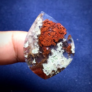 May include: A teardrop-shaped agate cabochon with a white, green, and brown base and a large, red-brown dendritic pattern.