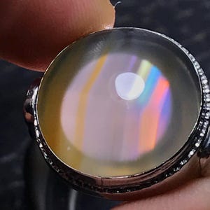 May include: A round, translucent cabochon gemstone set in a dark metal ring. The stone displays a smooth, milky surface with internal light refraction creating vertical bands of rainbow colors, including orange, yellow, and blue, against a pale base.