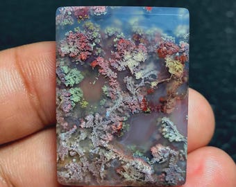 May include: A rectangular gemstone with a unique, colorful pattern. The stone features a mix of red, green, and purple hues, resembling a landscape or abstract design. The polished surface reflects light, highlighting the intricate details.