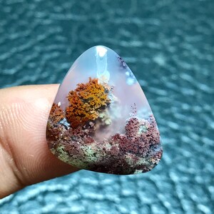 Op de afbeelding: Een driehoekige, doorschijnende agaat cabochon met een dendritisch patroon in tinten van bruin, oranje en wit. De agaat heeft een unieke, gelaagde uitstraling, die doet denken aan een landschap.