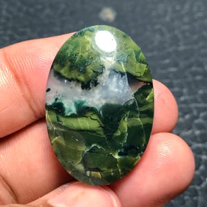 Puede incluir: Gema ovalada con un llamativo patrón verde y blanco. La superficie de la piedra presenta una mezcla de tonos verdes claros y oscuros, con zonas blancas y translúcidas. El acabado pulido refleja la luz, resaltando las texturas y variaciones naturales.