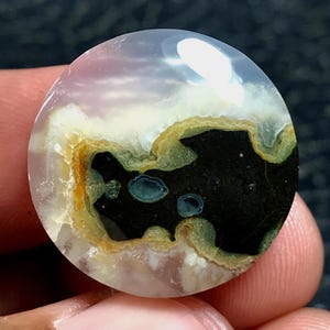 May include: A circular polished gemstone cabochon featuring a translucent pale lavender base with a central organic pattern of deep black, accented by bands of mustard yellow, olive green, and two small circular inclusions of teal blue.