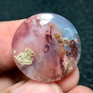 Peut inclure: Une pierre précieuse ronde et polie, présentant un mélange de couleurs rose, blanc et bleu. Des motifs complexes évoquent un paysage, avec des inclusions brunes et beiges. L'ensemble crée un effet naturel et organique.