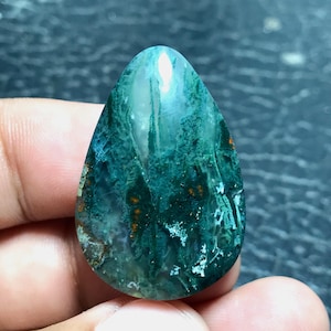 May include: A teardrop-shaped moss agate gemstone featuring a polished surface with deep forest green inclusions, translucent white areas, and small orange-brown mineral flecks throughout the stone, displaying a natural, organic pattern.