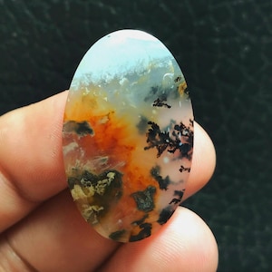 Peut inclure: Pierre précieuse de forme ovale avec un motif unique. La pierre présente un mélange de couleurs, notamment orange, noir et blanc, créant un design naturel et artistique. Le bijou est tenu dans une main, mettant en valeur sa taille et sa forme.