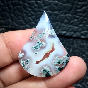 May include: A teardrop-shaped gemstone with a polished surface. The stone features a white base with swirling patterns of red and green inclusions. The top of the stone is pointed, and the overall shape is symmetrical.