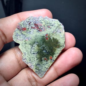 Könnte beinhalten: Ein Stück roher, ungeschnittener Stein mit strukturierter Oberfläche. Der Stein weist eine Mischung aus Farben auf, darunter hellgrün, grau und rot. Der Stein wird in einer Hand vor dunklem Hintergrund gehalten.