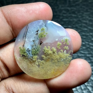 Pode incluir: Um cabochão redondo de ágata translúcido com uma base branca, rosa e cinza. A pedra tem um padrão único de dendritos verdes e marrons, que se assemelham a musgo ou líquenes.