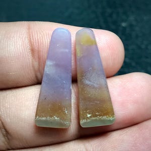 Puede incluir: Dos colgantes de piedras preciosas pulidas y alargadas. Los colgantes son una mezcla de tonos lavanda, marrón y amarillo. Los bordes inferiores tienen un tinte verde claro. Los colgantes se sostienen en una mano.