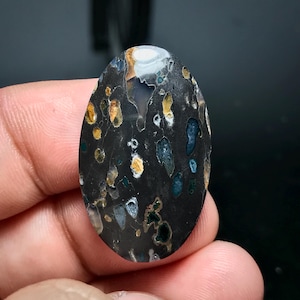 以下が含まれることがあります： 白、黄、青の斑点模様が施された、黒い楕円形のcabochonカットの宝石。石は磨かれた表面で、天然の宝石です。