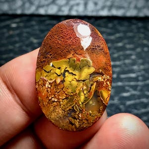 Op de afbeelding: Een ovaalvormige edelsteen met een mix van kleuren. De bovenste helft is roodbruin met een textuurpatroon, terwijl de onderste helft een levendig geel en goud patroon heeft. De steen wordt in een hand gehouden.