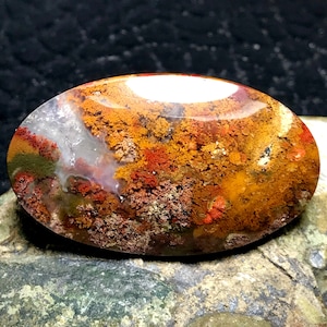 Puede incluir: Una piedra de ágata pulida de forma ovalada con un patrón único de dendritas rojas, naranjas y marrones. El ágata tiene una apariencia translúcida, permitiendo que la luz la atraviese.