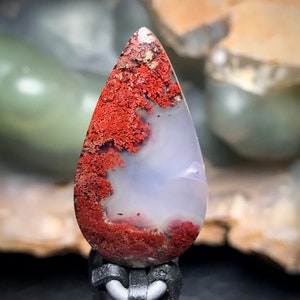 May include: A teardrop-shaped agate cabochon with a white base and red dendritic patterns. The agate is polished and has a smooth surface.