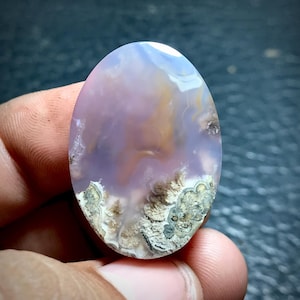 May include: A purple and white agate cabochon with brown and gray dendritic patterns. The cabochon is oval shaped and has a smooth, polished surface.