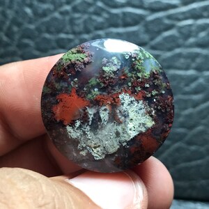 Pode incluir: Um cabochão de ágata redondo e polido com um padrão único de dendritos verdes, marrons e brancos. A pedra tem uma aparência translúcida e uma superfície lisa e polida.